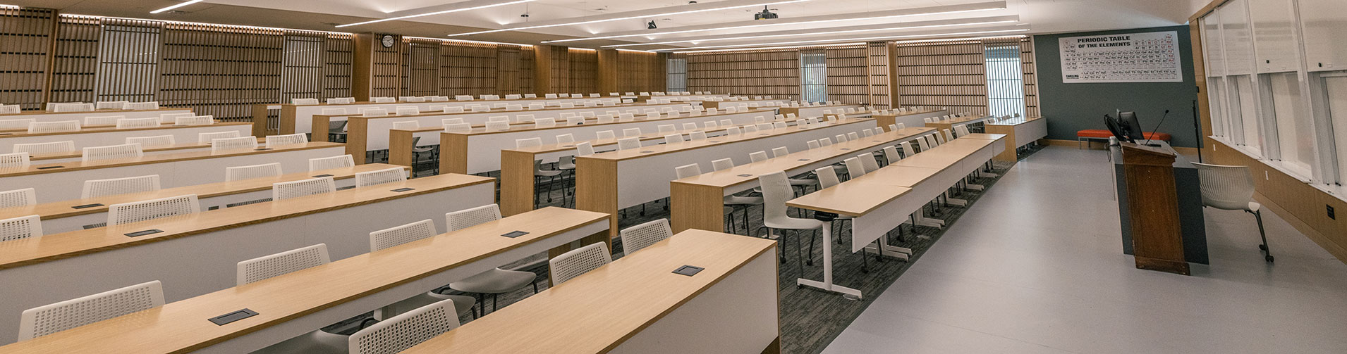 Empty modern lecture hall with rows of light wood desks, white chairs, and a dark brown podium.