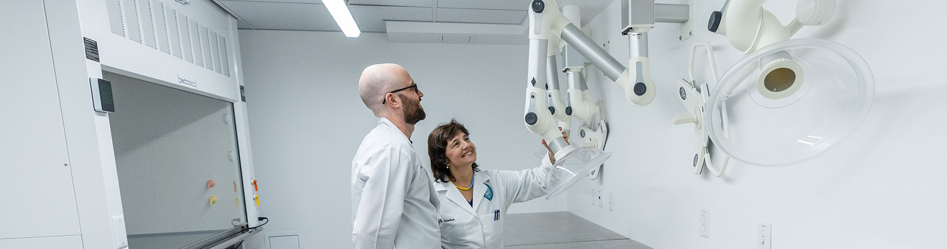 Two scientists in lab coats observe and adjust intricate lab equipment in a sterile room.