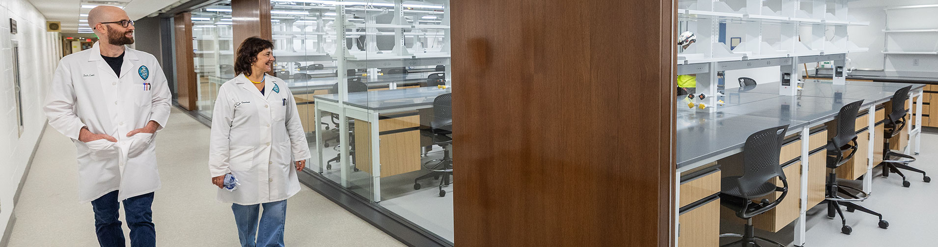 Two scientists in lab coats walk past a modern, empty laboratory.